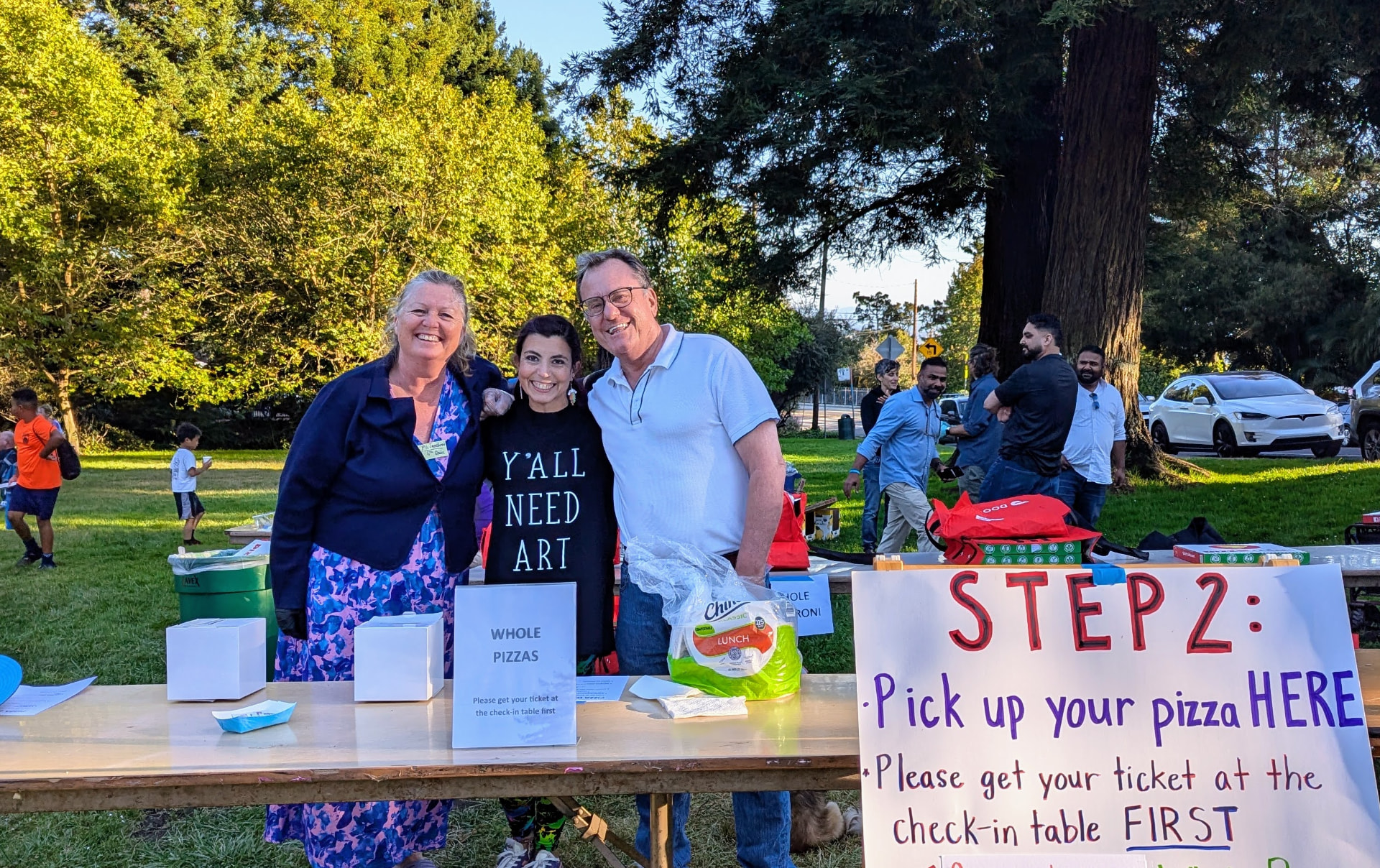 Three PTA volunteers smiling behind the pizza pickup table at the school's Pizza in the Park event