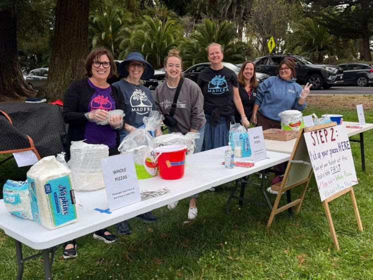 Six Madera volunteers in school shirts smile behind the pizza pickup tables at Pizza in the Park