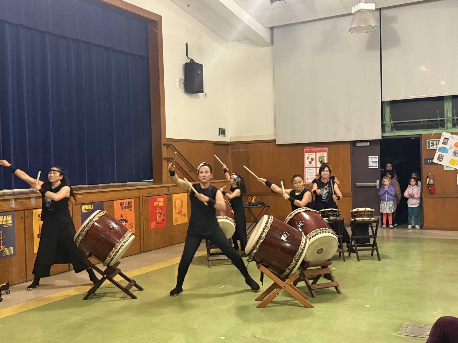 Taiko drummers in black perform on the Madera stage at Multicultural Night while families watch from the doorway